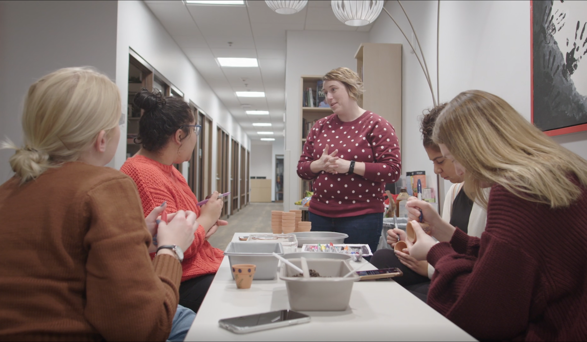 a woman stands next to a table where students sit, decorating small clay pots. The woman standing and one of the students sitting talk to each other.