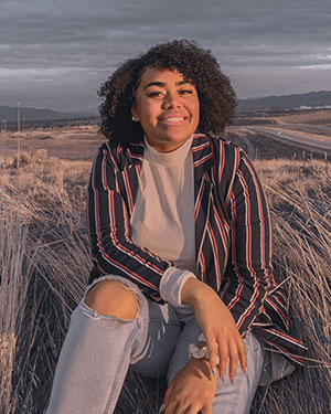 Olivia wears a striped blazer and torn jeans and smiles while sitting in a field of long grass