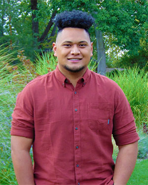 Will Satini wears a red button down shirt and smiles in front of green lush landscaping